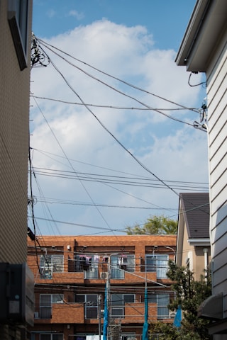 A residential area with a clear blue sky as the backdrop. The scene is framed by two buildings on each side with a row of apartment buildings in the background. Several telecommunication wires crisscross the upper portion of the frame. Clothes are hanging outside on the balconies of the apartments, drying in the open air.