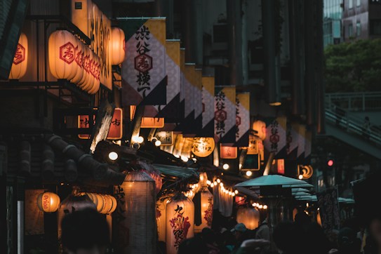 A dimly lit street adorned with traditional Japanese paper lanterns and banners, creating a warm and inviting atmosphere. The street is bustling with people, some of whom are partially visible beneath umbrellas. The architecture reflects a blend of traditional and modern elements, with the lanterns casting a soft glow against the darkening sky.