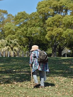 An elderly person wearing a plaid shirt and a hat is walking through a park using a walking cane. The person has a backpack and is surrounded by a grassy area with scattered fallen leaves. The background features tall, leafy trees and a clear blue sky.