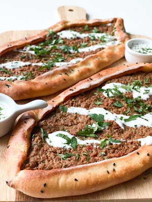 A wooden board holds two pieces of oval-shaped Turkish pide, topped with minced meat, herbs, and drizzled with a white sauce. Fresh parsley is scattered over the top for garnish. Small, round bowls, filled with a creamy sauce, accompany the pide on the board.