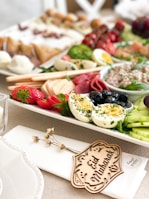 A beautifully arranged platter featuring a variety of foods including sliced hard-boiled eggs garnished with herbs, strawberries, cucumber slices, black olives, cured meats, and a spreadable paste. The platter is adorned with a small wooden piece inscribed with 'Eid Mubarak' placed on a napkin. The background is filled with an assortment of pastries and fresh greens.