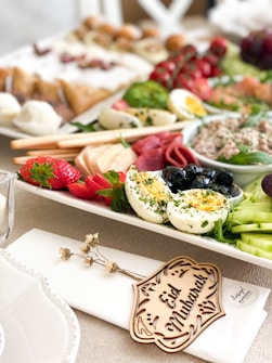 A beautifully arranged platter featuring a variety of foods including sliced hard-boiled eggs garnished with herbs, strawberries, cucumber slices, black olives, cured meats, and a spreadable paste. The platter is adorned with a small wooden piece inscribed with 'Eid Mubarak' placed on a napkin. The background is filled with an assortment of pastries and fresh greens.