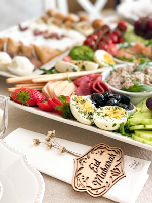 A beautifully arranged platter featuring a variety of foods including sliced hard-boiled eggs garnished with herbs, strawberries, cucumber slices, black olives, cured meats, and a spreadable paste. The platter is adorned with a small wooden piece inscribed with 'Eid Mubarak' placed on a napkin. The background is filled with an assortment of pastries and fresh greens.