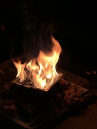 A small fire burns intensely on a square metal container with dried flowers or leaves arranged around it, set against a dark background.