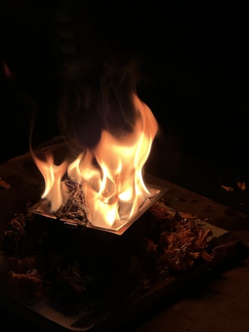 A small fire burns intensely on a square metal container with dried flowers or leaves arranged around it, set against a dark background.