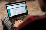 A person is using a Dell laptop, sitting on a textured carpeted floor. The individual is wearing ripped jeans and a dark jacket. The laptop screen displays an online form or questionnaire with options to select regarding gender and other information.