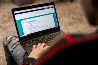 A person is using a Dell laptop, sitting on a textured carpeted floor. The individual is wearing ripped jeans and a dark jacket. The laptop screen displays an online form or questionnaire with options to select regarding gender and other information.
