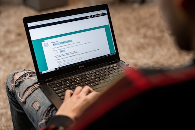 A person is using a Dell laptop, sitting on a textured carpeted floor. The individual is wearing ripped jeans and a dark jacket. The laptop screen displays an online form or questionnaire with options to select regarding gender and other information.