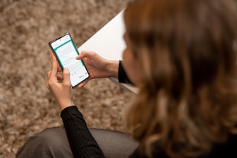 A person with light-colored hair is seated, holding a smartphone in both hands. The phone's screen displays a form or application interface with text and various icons. The person is wearing a dark sweater, and a white surface, possibly a table, is partially visible.