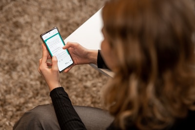 A person with light-colored hair is seated, holding a smartphone in both hands. The phone's screen displays a form or application interface with text and various icons. The person is wearing a dark sweater, and a white surface, possibly a table, is partially visible.