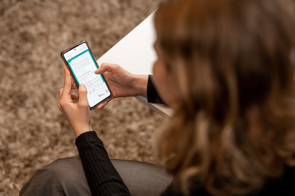A person with light-colored hair is seated, holding a smartphone in both hands. The phone's screen displays a form or application interface with text and various icons. The person is wearing a dark sweater, and a white surface, possibly a table, is partially visible.