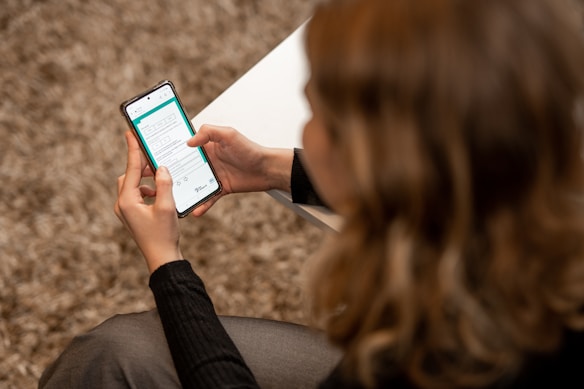 A person with light-colored hair is seated, holding a smartphone in both hands. The phone's screen displays a form or application interface with text and various icons. The person is wearing a dark sweater, and a white surface, possibly a table, is partially visible.