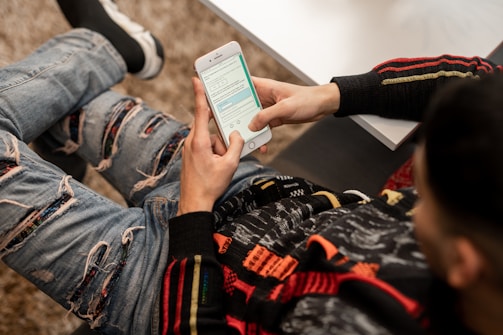 A person is sitting on a couch, wearing a colorful sweater and ripped jeans, holding a smartphone and engaging with a messaging app. The surrounding environment includes a table with a partially visible book or notebook.