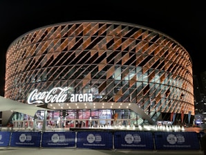 A large, modern arena with a distinctive rounded design featuring a complex pattern of illuminated panels. The facade displays the 'Coca-Cola arena' signage prominently. The venue is surrounded by barriers with 'World Tennis League' branding. The lighting creates a vibrant and inviting atmosphere at night, with lighting around the entrance and inside the building.