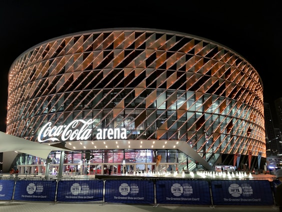 A large, modern arena with a distinctive rounded design featuring a complex pattern of illuminated panels. The facade displays the 'Coca-Cola arena' signage prominently. The venue is surrounded by barriers with 'World Tennis League' branding. The lighting creates a vibrant and inviting atmosphere at night, with lighting around the entrance and inside the building.
