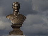 A bronze bust of a man wearing a decorated uniform with a medallion is set against a backdrop of dark, moody clouds. The sculpted figure appears formal and historical.