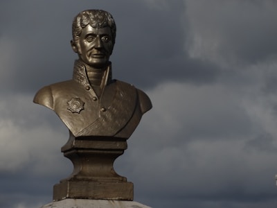 A bronze bust of a man wearing a decorated uniform with a medallion is set against a backdrop of dark, moody clouds. The sculpted figure appears formal and historical.