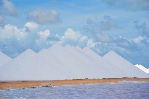Large, symmetrical white pyramids of salt stand in a line against a backdrop of a partly cloudy sky. The foreground features a body of water with a calm surface and a strip of sandy ground.