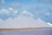 Large, symmetrical white pyramids of salt stand in a line against a backdrop of a partly cloudy sky. The foreground features a body of water with a calm surface and a strip of sandy ground.