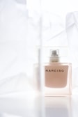 A glass bottle of perfume with the word 'NARCISO' on it, surrounded by soft, translucent fabric. The background is light and airy, contributing to a delicate and elegant presentation.