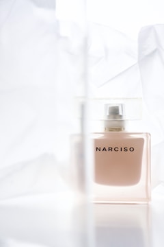 A glass bottle of perfume with the word 'NARCISO' on it, surrounded by soft, translucent fabric. The background is light and airy, contributing to a delicate and elegant presentation.