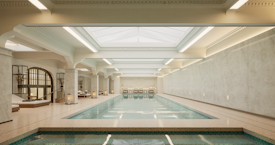 A luxurious indoor swimming pool with a serene and elegant design. The pool is surrounded by marble tiles and has a high ceiling with skylights allowing natural light to illuminate the space. Loungers and tables are positioned around the pool in a spacious, symmetrical arrangement. Ornate architectural details and muted colors create a sophisticated atmosphere.