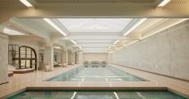 A luxurious indoor swimming pool with a serene and elegant design. The pool is surrounded by marble tiles and has a high ceiling with skylights allowing natural light to illuminate the space. Loungers and tables are positioned around the pool in a spacious, symmetrical arrangement. Ornate architectural details and muted colors create a sophisticated atmosphere.