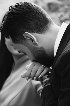 A black and white close-up image captures a man with a neatly trimmed beard kissing a woman's hand. The focus is on the intimate gesture, emphasizing the man's watch and the woman's rings. Their faces are partially visible, suggesting a romantic and tender moment.