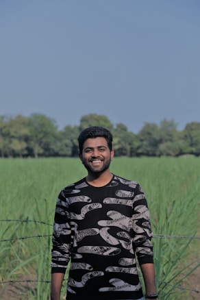 A person is standing in a field with tall green plants, smiling broadly. The sky is clear with a light blue hue, and a line of trees is visible in the background. The person is wearing a long-sleeved shirt with a graphic pattern, featuring the words 'Gardening' and other print designs. A barbed wire fence runs in front of the field.