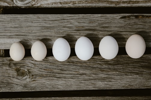 Six eggs are arranged in a line on a rustic wooden surface, showing a gradient of sizes.