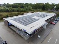 A large, flat-roofed commercial building with numerous solar panels installed on top, surrounded by a parking lot and green landscaping. There are several shopping carts lined up near the entrance, and a few cars parked nearby. The store's branding is visible on the facade.