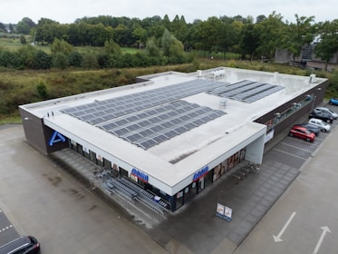 A large, flat-roofed commercial building with numerous solar panels installed on top, surrounded by a parking lot and green landscaping. There are several shopping carts lined up near the entrance, and a few cars parked nearby. The store's branding is visible on the facade.