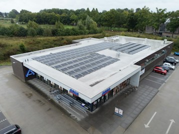 A large, flat-roofed commercial building with numerous solar panels installed on top, surrounded by a parking lot and green landscaping. There are several shopping carts lined up near the entrance, and a few cars parked nearby. The store's branding is visible on the facade.