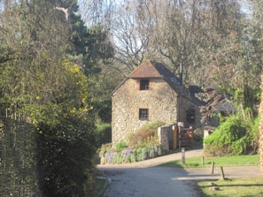 A rustic stone cottage is tucked away in a lush, wooded area. The cottage features a sloped roof and wooden window frames, surrounded by various trees and greenery. A dirt path leads up to the building, lined with purple flowers along the side. Sunlight filters through the leaves, creating a peaceful and serene atmosphere.