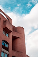A brick building with modern architectural design featuring curved and angular elements. The facade has several rectangular windows with reflections visible in the glass. Above, a clear blue sky is partially filled with white fluffy clouds. A bird is captured in mid-flight in the sky, adding a dynamic element to the scene.