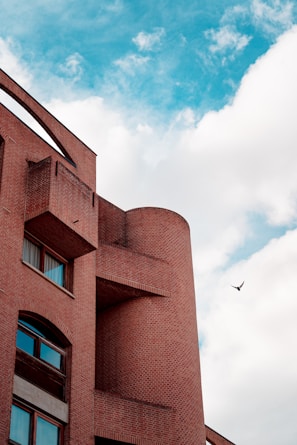 A brick building with modern architectural design featuring curved and angular elements. The facade has several rectangular windows with reflections visible in the glass. Above, a clear blue sky is partially filled with white fluffy clouds. A bird is captured in mid-flight in the sky, adding a dynamic element to the scene.