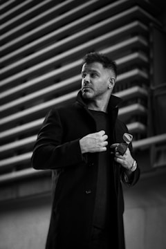 A man gazes thoughtfully to the side, standing in front of a modern architectural backdrop featuring horizontal lines. He is dressed in a dark coat, holding a pair of sunglasses in his hand.
