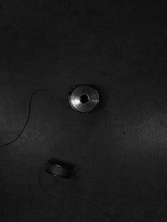 Two spools of thread rest on a dark, textured surface, connected by a strand of thread. The background is a muted, dark tone with subtle scratches and marks visible.