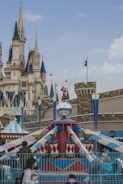A whimsical amusement park scene features a colorful carousel adorned with a mirrored disco ball and a playful character statue on top. In the background, a majestic castle with multiple spires and decorative flags adds to the enchanting atmosphere. Visitors are seen walking around, dressed in winter clothing.