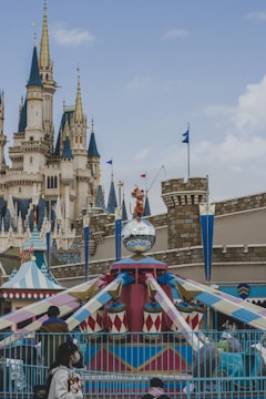 A whimsical amusement park scene features a colorful carousel adorned with a mirrored disco ball and a playful character statue on top. In the background, a majestic castle with multiple spires and decorative flags adds to the enchanting atmosphere. Visitors are seen walking around, dressed in winter clothing.
