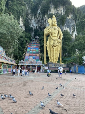 A large golden statue of a deity stands prominently against a backdrop of steep limestone cliffs. In the foreground, a wide plaza features several pigeons walking around while groups of people gather and take photos. A colorful staircase adorned with artistic details leads up to a temple entrance, surrounded by lush greenery.