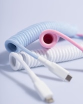 Three coiled USB cables in pastel colors, including blue, pink, and white, are arranged in a visually appealing manner. The cables are neatly coiled, displaying their colorful braided texture against a plain light background.