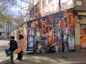 An urban street scene featuring vibrant graffiti art on a large wall, depicting a neon-lit alley reminiscent of a bustling cityscape. Two people are nearby, one holding a skateboard with orange accents, while the other looks at a phone. Scaffolding is visible in the background, and scattered leaves cover the pavement.