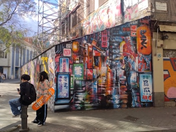 An urban street scene featuring vibrant graffiti art on a large wall, depicting a neon-lit alley reminiscent of a bustling cityscape. Two people are nearby, one holding a skateboard with orange accents, while the other looks at a phone. Scaffolding is visible in the background, and scattered leaves cover the pavement.