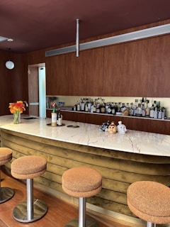 A modern bar setup features a sleek marble countertop with brown textured stools. Behind the bar, a wall showcases a variety of liquor bottles. Decorative items include two intricately designed skulls and a vase with vibrant orange flowers. The wooden cabinetry adds warmth to the space.