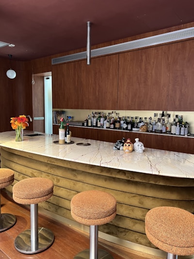 A modern bar setup features a sleek marble countertop with brown textured stools. Behind the bar, a wall showcases a variety of liquor bottles. Decorative items include two intricately designed skulls and a vase with vibrant orange flowers. The wooden cabinetry adds warmth to the space.