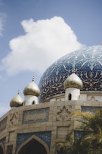 A large, intricately designed mosque dome with golden spires and blue and gold detailed patterns. The dome is surrounded by smaller golden domes, and the architecture is adorned with geometric and floral designs. A partly cloudy sky serves as the backdrop.