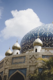 A large, intricately designed mosque dome with golden spires and blue and gold detailed patterns. The dome is surrounded by smaller golden domes, and the architecture is adorned with geometric and floral designs. A partly cloudy sky serves as the backdrop.