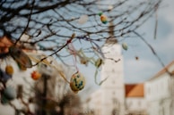 Branches decorated with colorful, painted Easter eggs hang delicately in the foreground, with a building featuring a tall tower slightly out of focus in the background. The eggs are adorned with various patterns and hang from ribbons.
