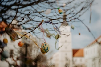 Branches decorated with colorful, painted Easter eggs hang delicately in the foreground, with a building featuring a tall tower slightly out of focus in the background. The eggs are adorned with various patterns and hang from ribbons.